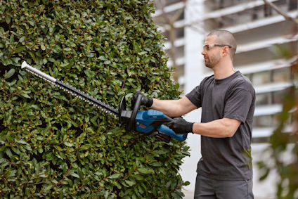Man trimmar buske med trådlös häcksax, klädd i grå t-shirt och skyddsglasögon.