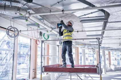 En person i skyddsväst och hjälm arbetar med borrmaskin på en byggarbetsplats inomhus.