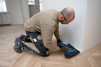 Person installerar golvlister på trägolv med elverktyg i ett rum.