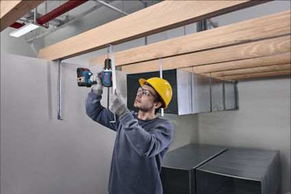 Person med gul hjälm och skyddsglasögon borrar i metallram under träbjälkar inomhus.