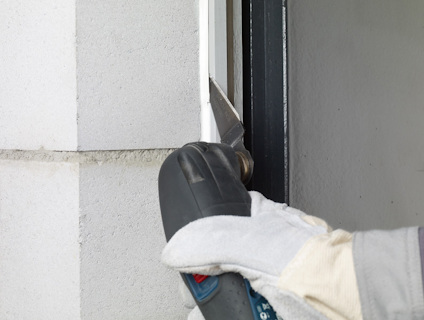 Hand med arbetshandske använder ett elektriskt multiverktyg för att såga i en dörrkarm av trä.