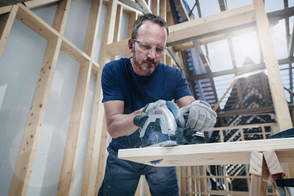 Person som använder en cirkelsåg för att såga trä, med skyddsglasögon och hörselskydd, omgiven av en träkonstruktion.