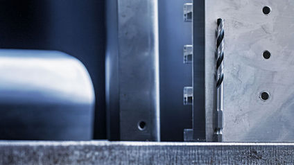 Närbild av stålplattor med borrade hål och en utskjutande spiralborr på en slät yta.