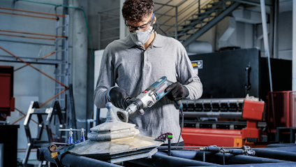 En person slipar en metallplatta med en elektrisk vinkelslip i en industrimiljö, bär skyddsglasögon och mask.