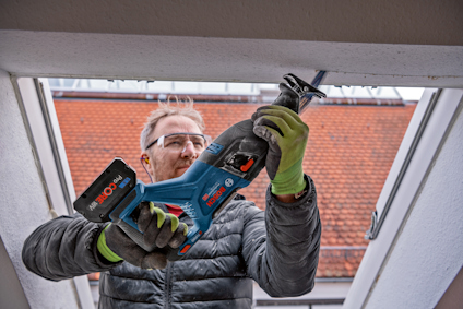 En person använder en blå elektrisk sticksåg med ett Bosch-batteri i ett träramförsett öppet fönster.