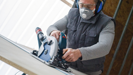 Person with goggles and mask using a circular saw on a wooden board.