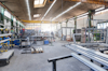 Verkstad med metallstänger, verktyg och industriutrustning. Takbelysning och stora fönster syns i bakgrunden.