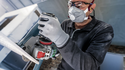 Person med skyddsglasögon och ansiktsmask använder en slipmaskin på metallplåt.