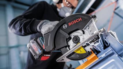 Person som använder en Bosch cirkelsåg på metallstruktur, med skyddshandskar och hörselskydd.