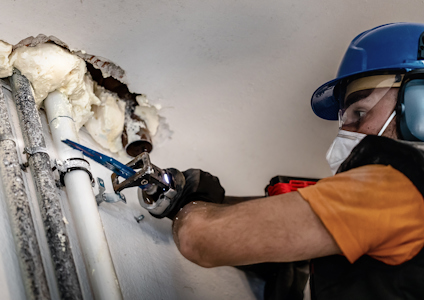 En person installerar rör i en vägg; skumplast och verktyg är synliga. Personen bär hjälm och ansiktsskydd.
