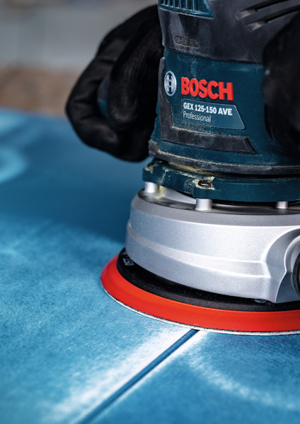 Två händer håller en Bosch GEX 125-150 AVE Professional excenterslip på en blå yta.