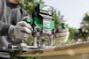 Person using a green and black electric router on a wooden plank, wearing gray gloves.