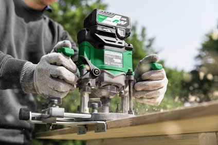 Person using a green and black electric router on a wooden plank, wearing gray gloves.