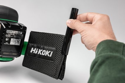 En hand håller en penna bredvid verktygsförvaringsväska med texten "Hikoki". Verktyg syns delvis i bakgrunden.