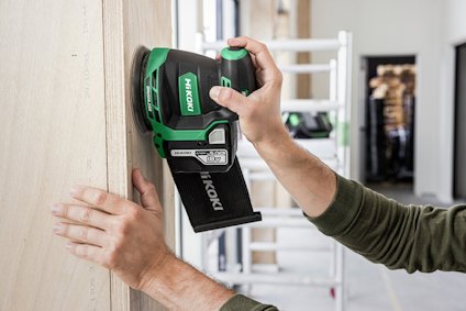 En person använder en grön HiKOKI-slipmaskin på en trädörr eller skiva.