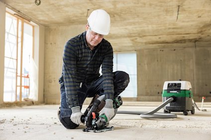 Person med hjälm och handskar arbetar på ett byggprojekt, använder ett verktyg på ett betonggolv, dammsugare i bakgrunden.