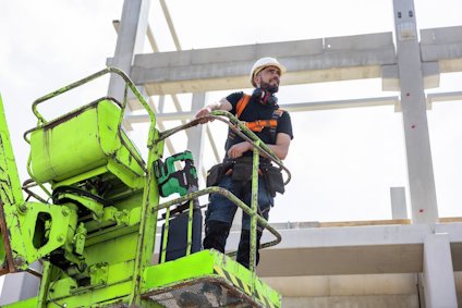 En byggarbetare i skyddskläder står på en grön arbetsplattform framför en konstruktion av betongbalkar.