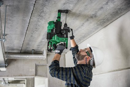 Person i hjälm och skyddsglasögon borrar i ett betongtak med en grön elektrisk borrmaskin.