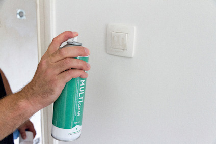 En hand håller en grön sprayburk märkt "MULTICLEAN" nära en vit vägg med en vit strömbrytare.