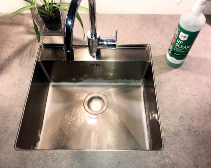 Rostfri diskho med kran, växt i hörn och flaska märkt "HP CLEAN" på en bänkskiva.