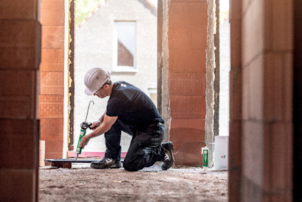 Person som arbetar på ett byggprojekt, iförd skyddshjälm och knäskydd, använder ett verktyg parallellt med murade väggar.