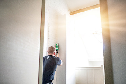 En person applicerar tätning med en caulk pistol på en vägg nära ett fönster med starkt dagsljus.