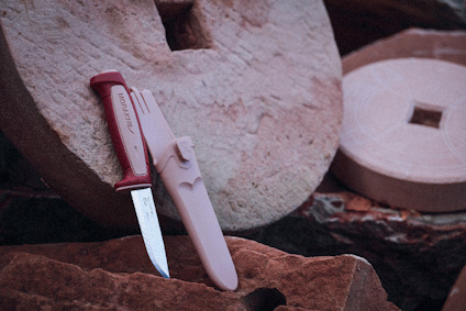 Kniv med röd handtag och beige hölster, placerad på en rund, ojämn sten.
